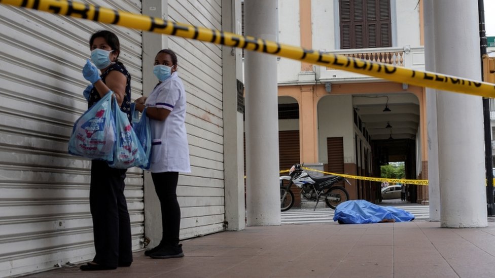 Two women standing next to a closed store in Guayaquil and a covered dead body lying on the sidewalk in the background